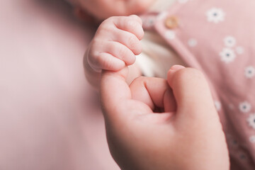 Mother holds newborn hand on blurred background at home. Woman and baby girl spend time together in bedroom filled with sunlight closeup