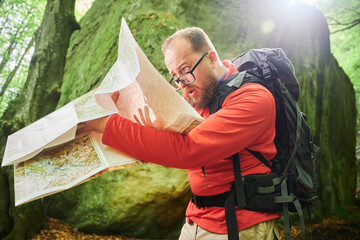Bearded tourist man looks puzzled, studies map while stands in front of large rock in dense forest....