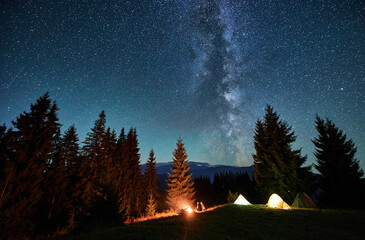 Fototapeta premium Night camping in mountains. Amidst forested mountain landscape, two tents glow warmly beside campfire under brilliant, star-filled sky. Silhouettes of tourists resting at campsite under Milky Way.