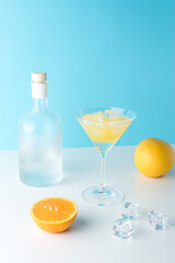 Tropical cocktail in martini glass with ice cubes and bottle of alcohol on white table. Minimal summer drink composition.