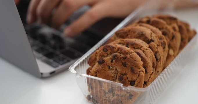 A businesswoman typing on a laptop and then taking a chocolate chip cookie from a box of other ones and getting ready to eat it during a break at work. Close up.
