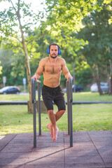 Shirtless athletic man listening music and training outdoors on parallel bars