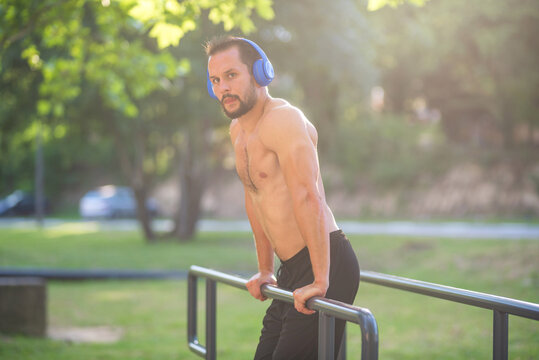 Young handsome man listening to music and exercising outdoors on parallel bars in the park