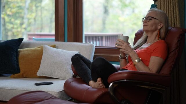 Mature woman with gray hair sits in a red leather recliner, wearing an orange shirt, black pants, and glasses. She is drinking from a white mug, with a phone resting on the couch beside her.