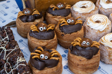 Close-up of festive pastries on a tray, likely donuts, decorated with chocolate icing and pretzel spiders for Halloween. Elaborate meringue swirls and a chocolate brownie are also visible.