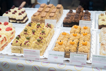 High-angle close-up of a display of cakes on white trays. Flavors include pistachio with buttercream, pumpkin spice with decorative toppers, and rich chocolate. Price tags are visible for each cake.
