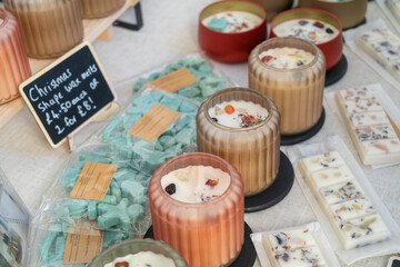 Close-up of colorful candles and wax melts for sale at a market stall. Small round candles in clear or frosted containers, with blocks of cream wax melts and a sign for Christmas-shaped melts.