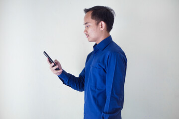 Smiling young Asian businessman in a blue shirt with a happy, surprised and happy expression while holding his smart phone on a white background. Ideal for technology, communication, social media enga