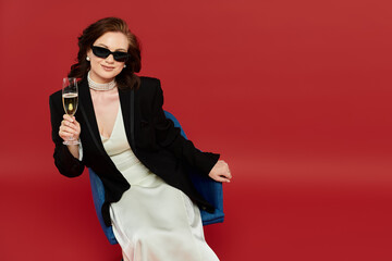 A young beautiful woman in elegant attire enjoys a glass of champagne while posing confidently.