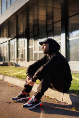 Young african american man in baseball cap sitting on border on urban street