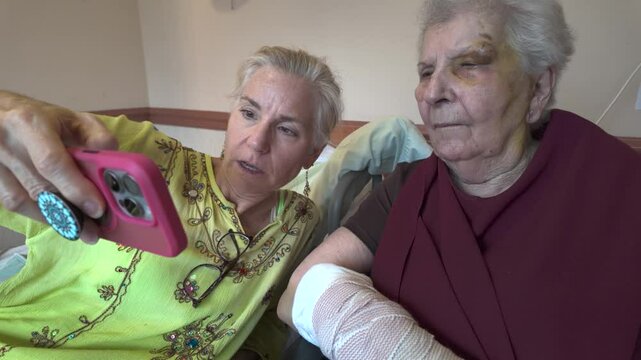 A woman in a green shirt is holding up a pink smartphone to take a picture of her elderly mother who is wearing a burgundy shirt and has a bandage on her arm.