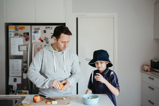 man cutting fruits on the kitchen counter with young boy beside him getting ready for school