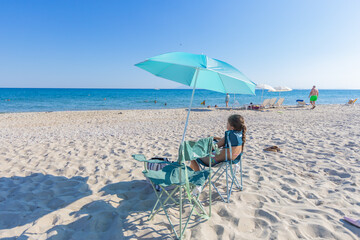 Child sitting on beach chair under sunshade on summer vacation.