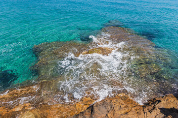 Crystal-clear waters gently lap over rocky formations at the shore of a beach in Greece.