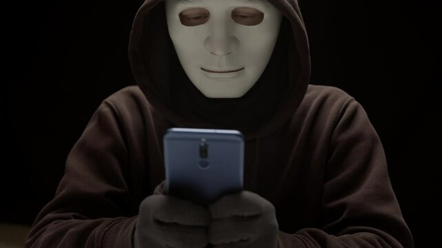 A hacker in a mask holds a phone in his hands. Cybersecurity concept and financial fraud using a smartphone.