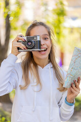 Caucasian young woman tourist traveling on weekends exploring town looking city paper map, making photo pictures on retro vintage camera outdoors. Happy teenager girl traveler on urban city street.