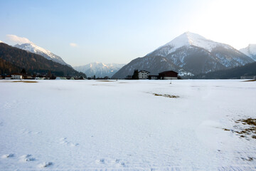 A snow-covered field behind which the houses of the village and small mountains are covered with forest and snow. Picturesque winter landscape