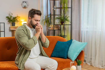 Tensed young man sitting on sofa at home folding palms together pray ask for help, thinks feel...