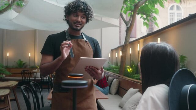Friendly restaurant worker guy Arabian Indian male waiter in apron serve woman client in cafe customer service smiling man serving in cafeteria communicate with visitor write food order digital tablet