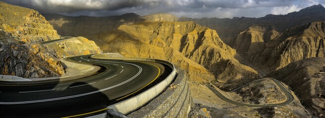Winding mountain road with dramatic shadows and rugged terrain under a cloudy sky