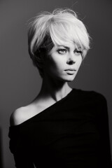 Stylish young woman with short blonde hair in a black off-shoulder dress, expressing a contemplative mood against a dark, soft-focus background.