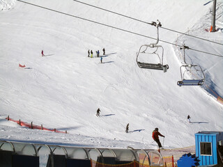 Skiers descend on a wide winter ski track. The empty benches of the suspended cable car move above them. Winter sports and recreation