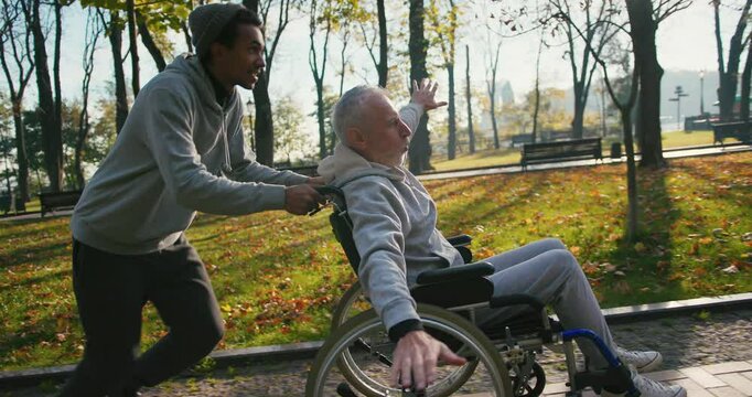 Hispanic man taking care of old disabled friend in wheelchair. Friends running, having fun together in autumn park. Healthcare, disability, mobility, friendship