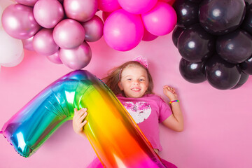 Portrait of a happy little girl lying on a pink background with balloons.  and holding the number 7. Happy birthday. Pink background.