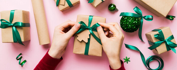 Fototapeta premium Woman is packing surprise gift for Christmas holidays, sitting at workplace with craft tools, top view. Woman wrapping Christmas gift at colored table