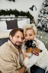 A couple enjoys a warm embrace with their dog, surrounded by holiday decorations at home.