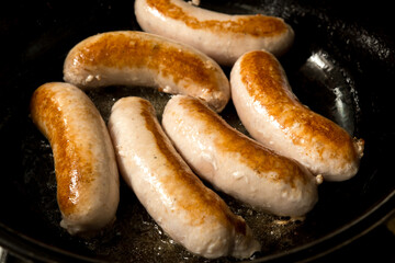 Six light-colored sausages frying in a pan with oil. They are in various stages of browning, suggesting they are being cooked on medium to high heat. Perfect for cooking and food photography themes