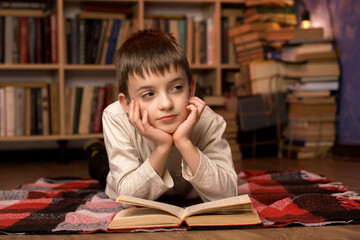 Thoughtful boy lying on a plaid blanket in a cozy library, daydreaming with an open book,...