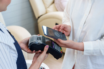 Senior Asian woman uses smartphone for contactless payment in nail spa, interacting with nail technician holding the card reader. Transaction ease and security of mobile payments in a professional