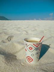 A cup coffee with a straw resting on sandy beach under a clear blue sky creating a calm summer scene in Camiguin Island in the Philippines