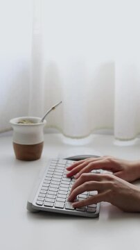 A south american freelancer is working from home in a minimal environment lit by natural light. She is typing on a keyboard, clicking the mouse and drinking yerba mate infusion.