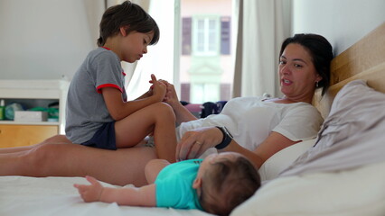 Obraz premium Mother lying on a bed with her baby and young boy, playfully engaging in a warm family moment, with the baby resting comfortably in a relaxed home environment