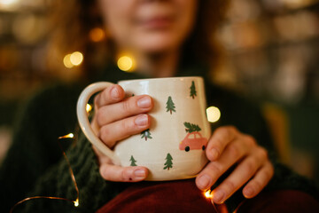 Festive close-up of hands holding a Christmas mug, warm holiday atmosphere. Close-up of woman hands holding a holiday mug, with bokeh lights creating a Christmas atmosphere