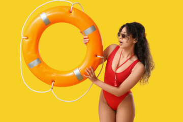 Beautiful young shocked African-American female lifeguard with ring buoy on yellow background
