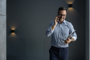 Smiling Businessman Talking On Phone Holding Laptop Indoors