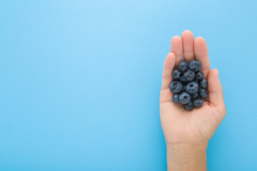 Young adult woman opened palm holding heap of fresh blueberries on light blue table background. Pastel color. Closeup. Empty place for text. Top down view.