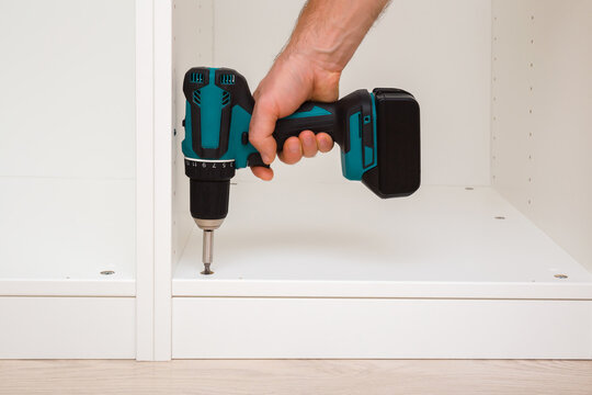 Young adult man hand using professional battery screwdriver and screwing screw in white board of base cabinet frame. Assembling new home wooden furniture. Closeup. Front view.