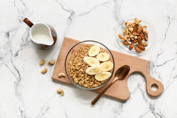 Wooden cutting board, nuts, bowl with tasty granola and banana on marble background