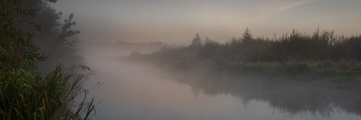 damp foggy twilight over the river with marshy densely overgrown banks. beautiful widescreen panoramic side view in 15x5 format