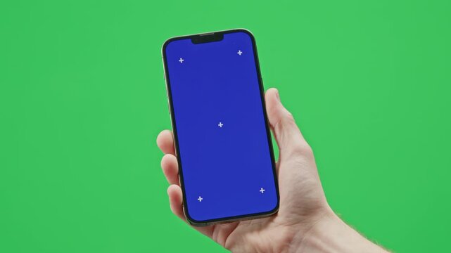 A modern smartphone is held in a man&rsquo;s hand against a green background. The phone of the screen has a chromakey blue image with tracking markers.