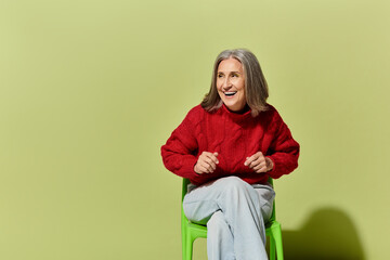 Fototapeta premium A radiant mature woman with grey hair smiles while dressed in a cozy red sweater against a vibrant green wall.