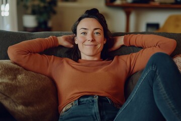 Fototapeta premium Happy middle aged woman in her 40s sitting on sofa in living room at home. Smiling beautiful mature lady relaxing on couch with candid pose in cozy house.