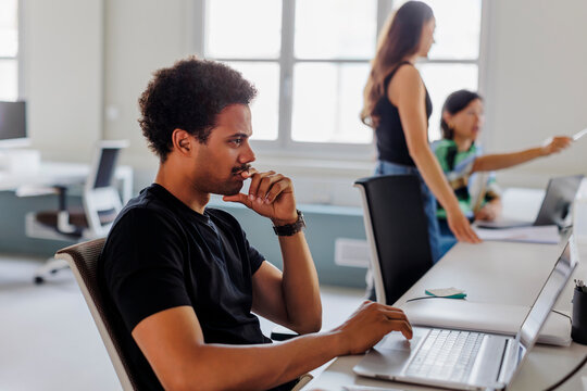 Thoughtful male developer working on laptop in busy office