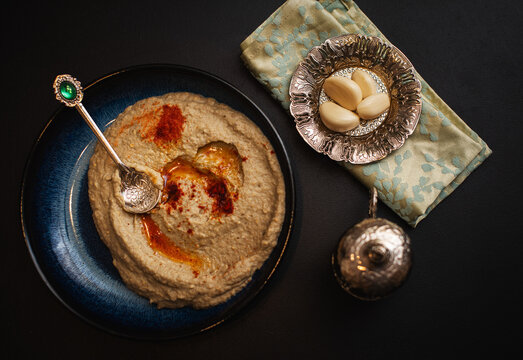 Close-up, top view. Homemade Baba Ganush on a blue plate surrounded by silverware. Black background.