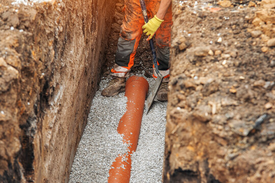 Construction worker installing drainage pipe in a trench during daylight, surrounded by gravel and soil