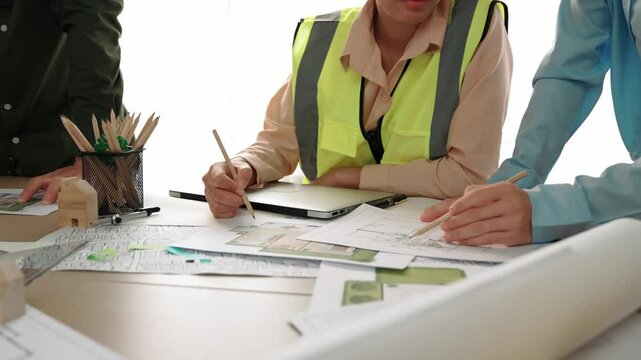 Smart architect and safety engineer discussion about building blueprint on table with architectural document, measurement tools and wooden block scatter around at modern office. Closeup. Delineation.